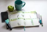A planner with a green mug and granny smith apple above it, pens on the side of it and a cell phone on the left.